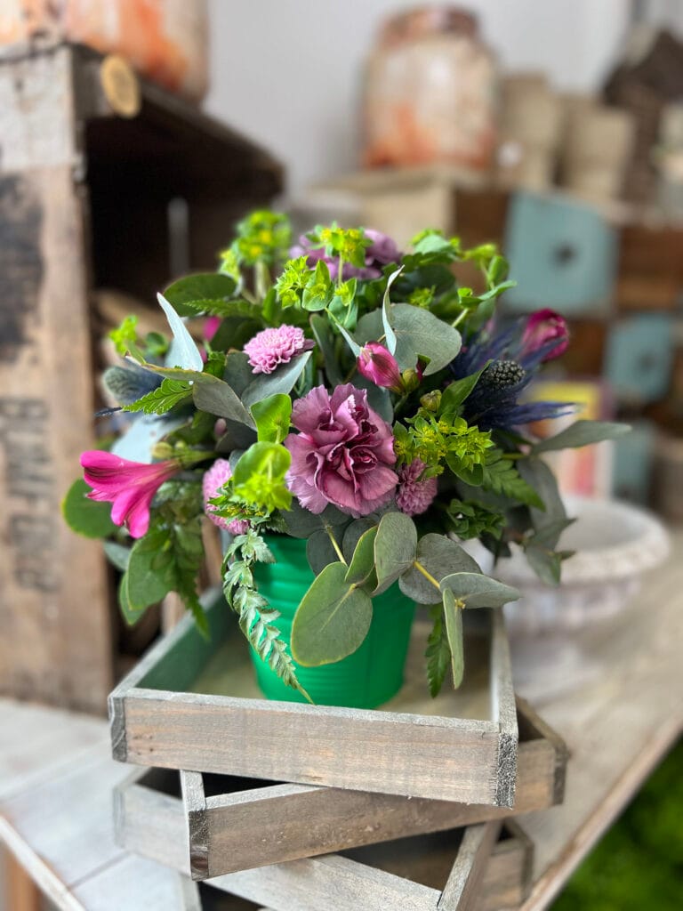 Colourful flower arrangement in green pot on wooden tray.